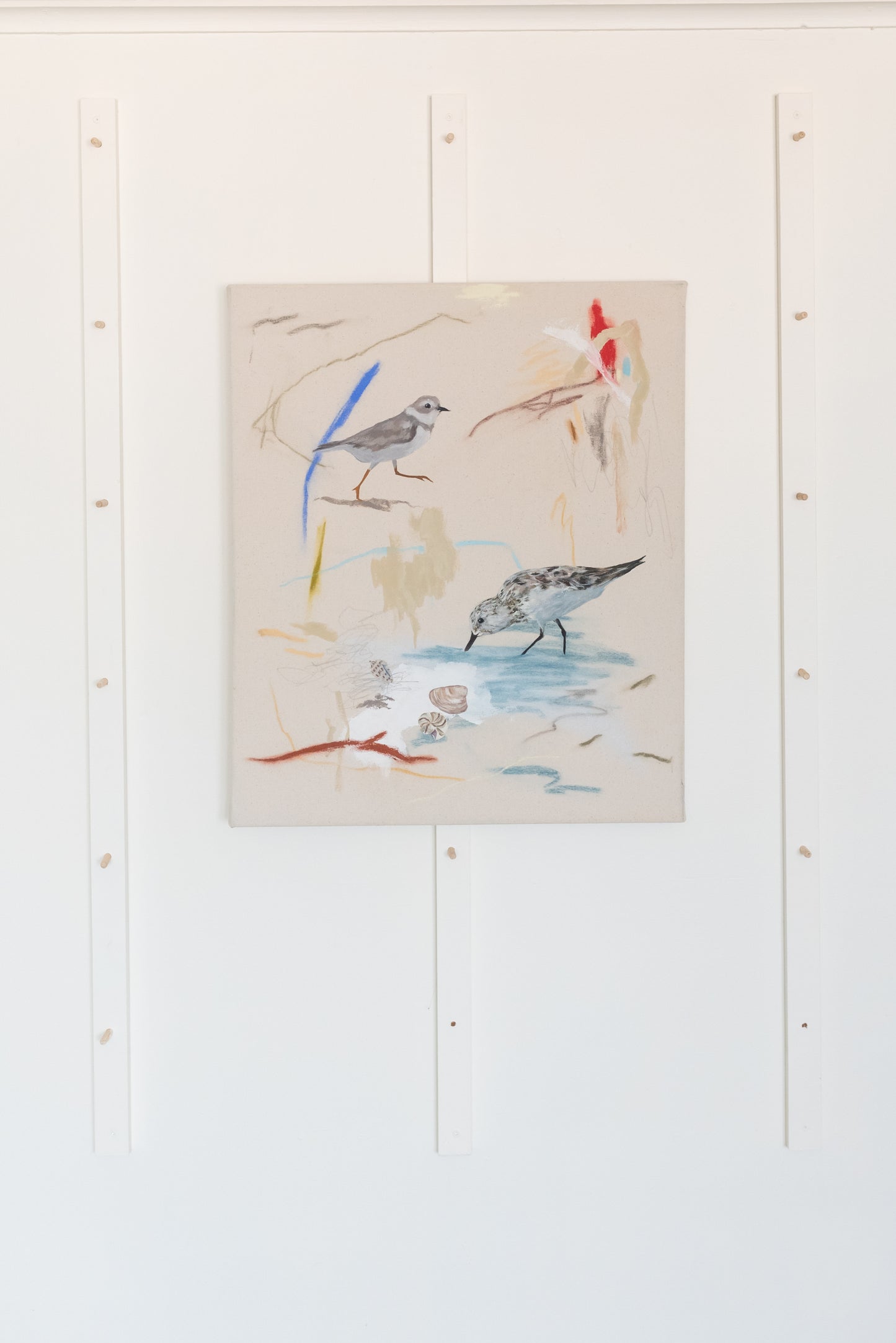 Coastal-inspired painting featuring a piping plover, sandpiper, and various seashells, displayed in a minimalist studio setting. The natural beach tones of the painting contrast beautifully against the white wall.