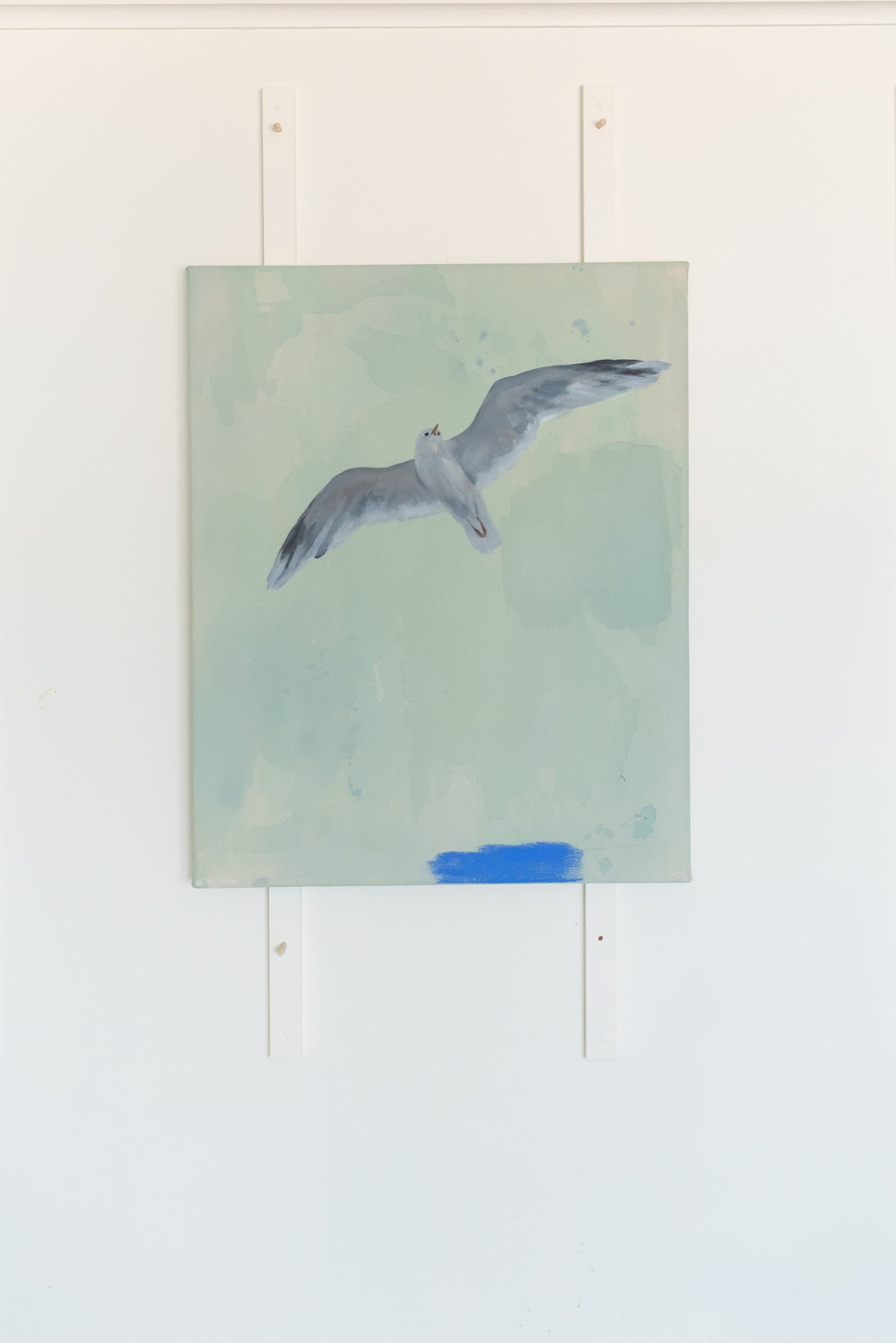 Coastal-inspired acrylic painting featuring a seagull in flight, displayed on a white studio wall in Stevie Sisson's Nova Scotia studio. The artwork's light pastel tones contrast against the minimalist setting, highlighting its calming beach theme.
