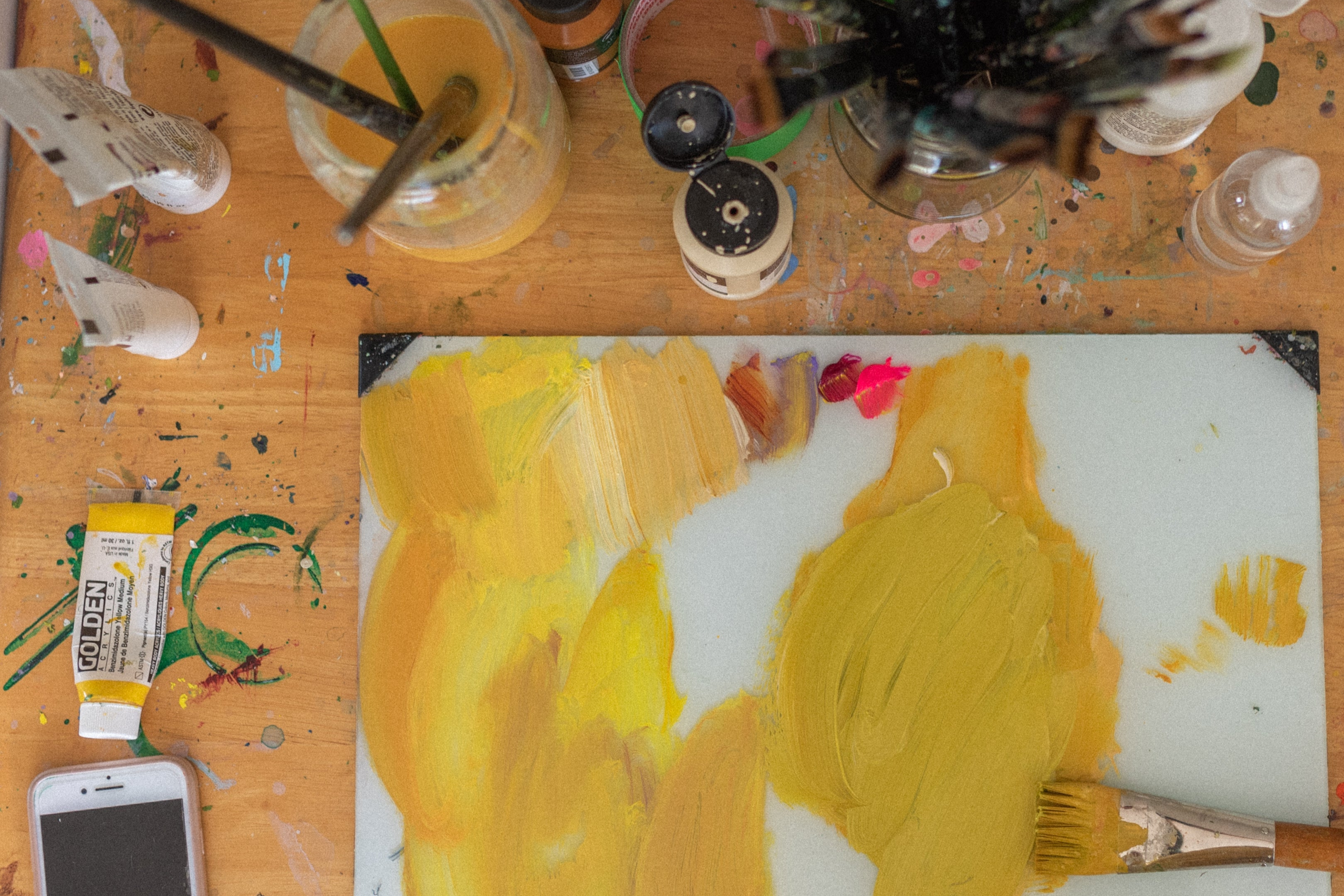Artist Stevie Sisson's workspace with a glass palette featuring vibrant yellow and ochre paint, surrounded by paintbrushes, art supplies, and a smartphone on a cluttered wooden table.