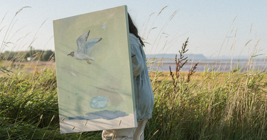 Artist Stevie Sisson holding a large canvas painting of a seagull while standing in a field of tall grasses near the coast, with the ocean visible in the background.