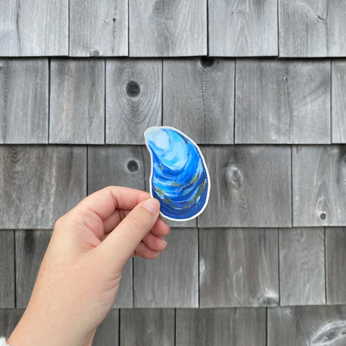 A hand holding a die-cut blue mussel shell sticker against a weathered gray wooden shingle background.