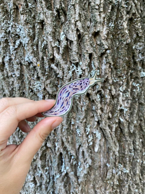 Hand holding a leopard slug sticker against a tree bark background.