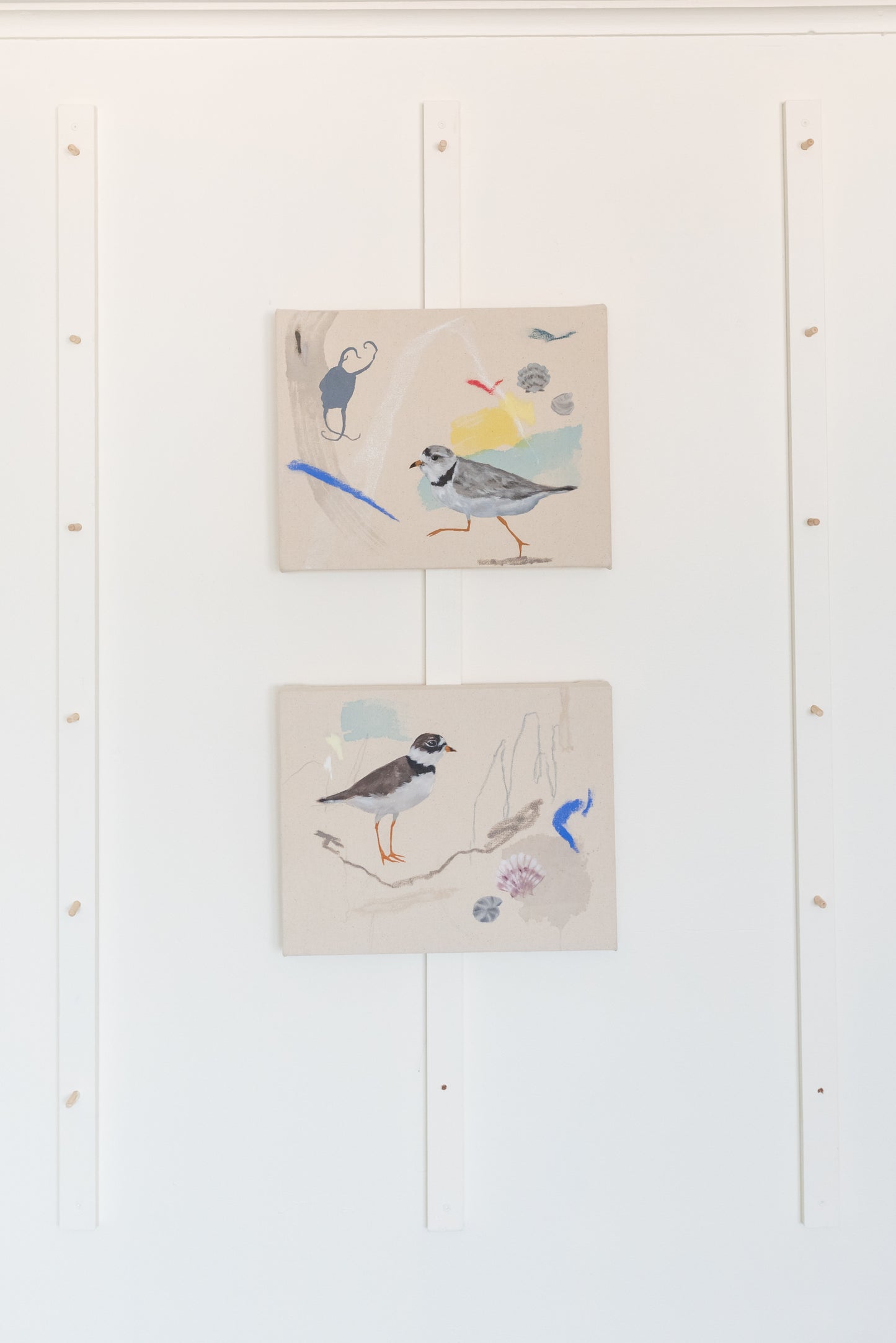 Two coastal-inspired paintings featuring a piping plover standing on the shore, displayed in Stevie Sisson's minimalist studio setting. The natural beach tones of the painting contrast beautifully against the white wall.